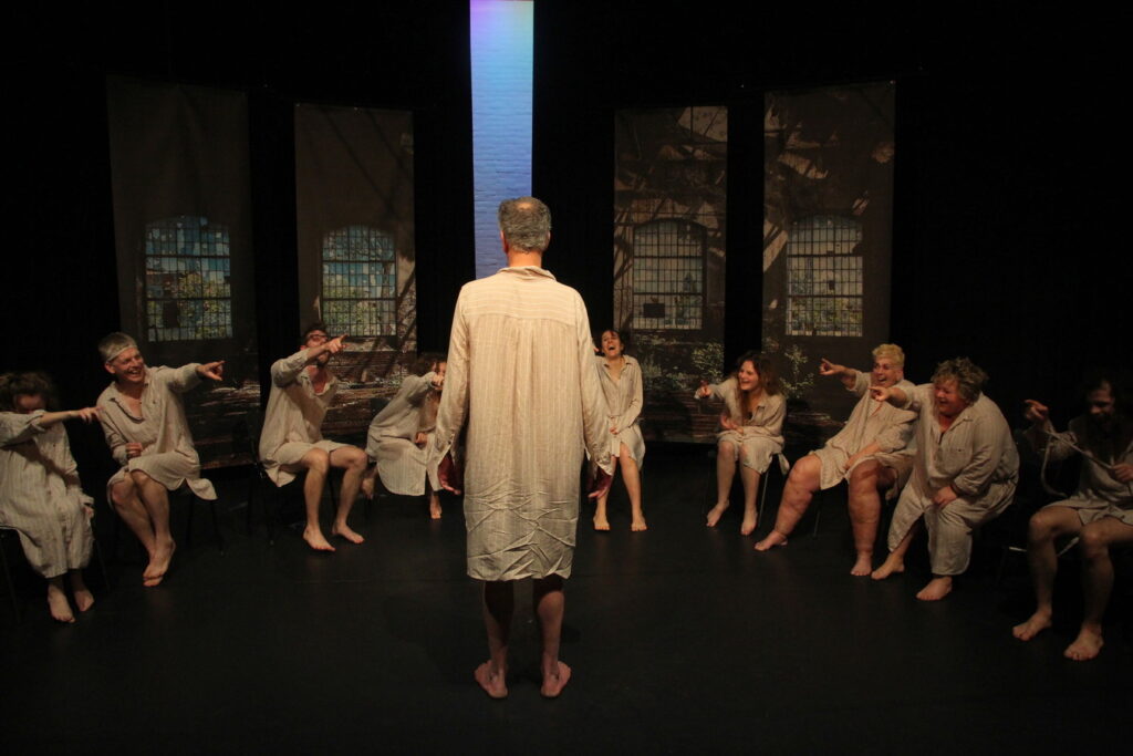 Een groep mensen in witte gewaden zit in een halve cirkel op stoelen en wijst naar een persoon in het midden die met de rug naar de camera staat. Op de achtergrond zijn grote ramen met metalen roosters zichtbaar, met een verticale lichtstraal die door het midden van de ruimte schijnt.